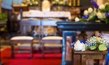 Flower arrangement sits on a wooden pew; in the background, padded kneelers face an adorned altar with floral decorations, inside a softly lit and ornate church interior.