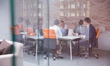 A group of six people sit around a conference table, engaging in discussion, with laptops in a modern office featuring large windows and orange-accented chairs.