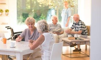 Elderly individuals are seated around a well-lit living room, engaging in conversation and activities. In the background, a caregiver in a blue uniform stands by the window.